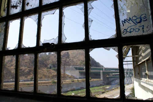 View through a broken window, showcasing a highway and hillside in the background, with graffiti on the glass.