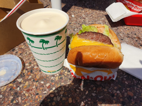A cheeseburger with lettuce and a drink in a cup on a table, with a takeout box in the background.