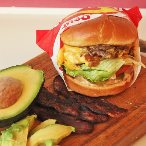 A juicy cheeseburger with lettuce and tomato, served with avocado slices and crispy bacon on a wooden cutting board.