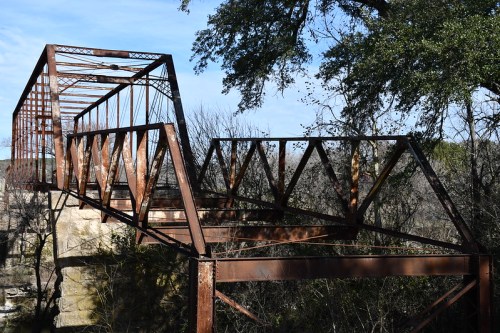 Rusty metal bridge frame overgrown with vegetation against a clear blue sky.