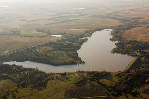 Aerial view of a winding lake surrounded by lush green fields and trees under a hazy sky.