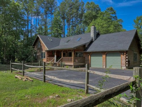 A rustic log cabin surrounded by trees, featuring a porch, garage, and a spacious driveway.