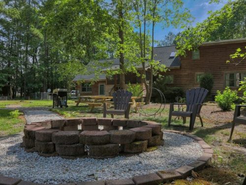 A cozy outdoor space featuring a stone fire pit, chairs, and a wooden cabin surrounded by trees and greenery.