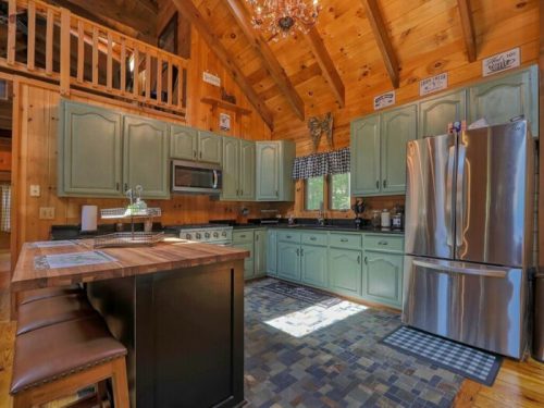 Cozy wooden kitchen with green cabinets, stainless steel appliances, and a rustic charm, featuring a large island and chandelier.