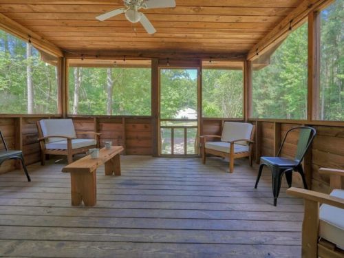 A cozy screened porch with wooden walls, a ceiling fan, and seating arranged around a central wooden table.