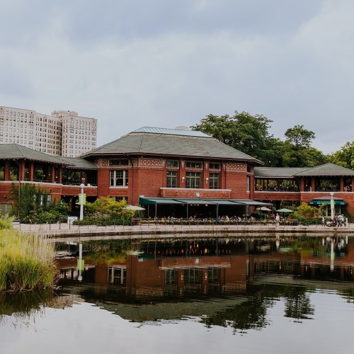 A brick building with a green roof reflects in a calm lake, surrounded by lush greenery and a cloudy sky.