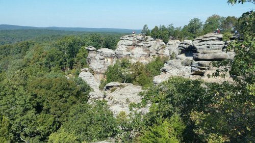 Rocky cliffs surrounded by lush green trees under a clear blue sky, with people exploring the area.