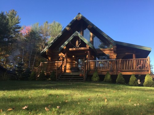A cozy log cabin with a green roof, surrounded by trees and a grassy lawn under a clear blue sky.