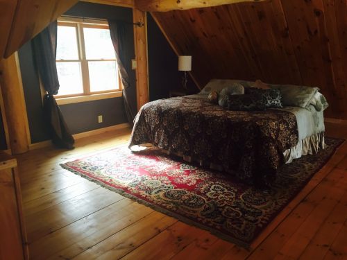 Cozy attic bedroom with a wooden ceiling, a large bed, and a patterned rug, illuminated by natural light from a window.