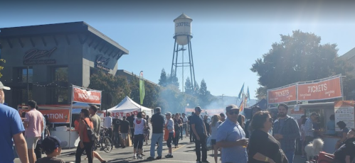 A lively outdoor event with people, tents, and a water tower in the background under a clear blue sky.