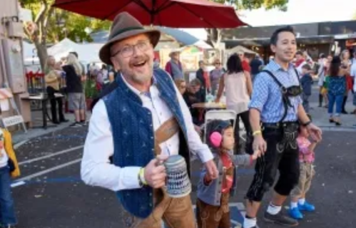 A man in traditional attire holds a beer mug, smiling, while a child and another man in lederhosen walk beside him at a festival.