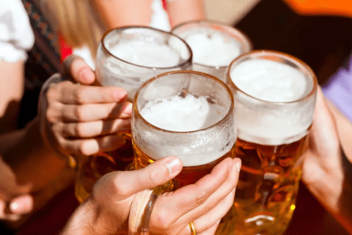 Four hands holding large beer mugs, clinking together in celebration, with frothy beer visible in each glass.