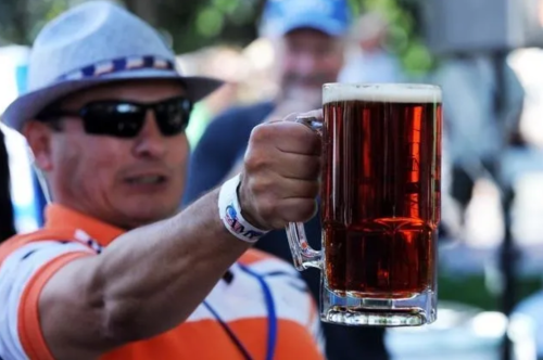 A man in sunglasses and a hat raises a large mug of dark beer, smiling at a gathering.