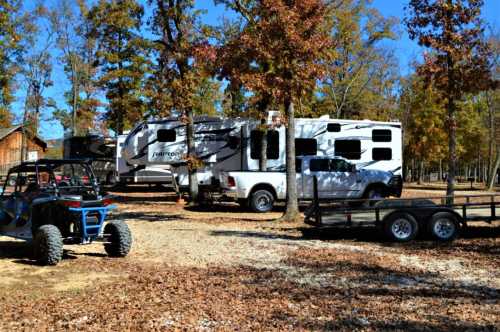 A white RV parked among trees with autumn leaves, alongside a truck and a trailer in a wooded area.