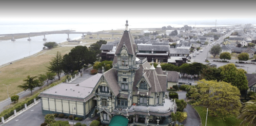 Aerial view of a large, ornate Victorian mansion near a waterfront, surrounded by greenery and a small town.