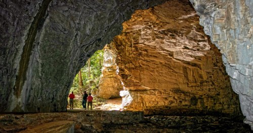 Two hikers explore a large cave with illuminated rock formations and greenery visible in the background.