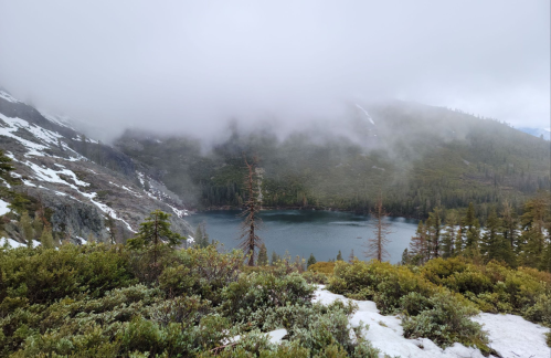 A serene lake surrounded by misty mountains and lush greenery, with patches of snow on the ground.