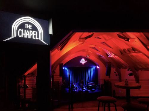A dimly lit music venue with a sign reading "The Chapel," featuring colorful lighting and a stage setup.