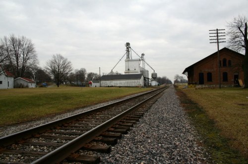 A view of a railway track leading to a grain elevator and an old brick building, set against a cloudy sky.