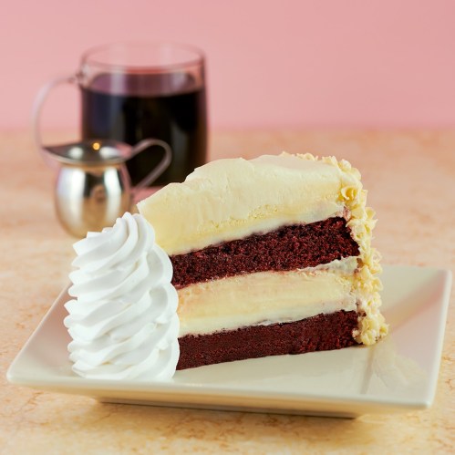 A slice of red velvet cake with cream cheese frosting and a dollop of whipped cream, served on a plate with coffee.