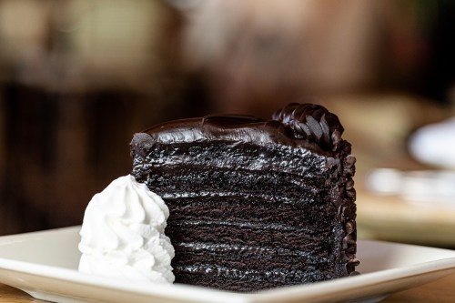 A slice of rich chocolate cake with multiple layers, topped with dark frosting and served with a dollop of whipped cream.