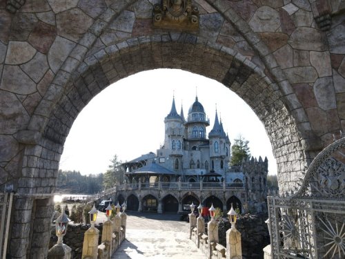 A grand stone castle with towers, viewed through an ornate archway, surrounded by trees and a clear sky.