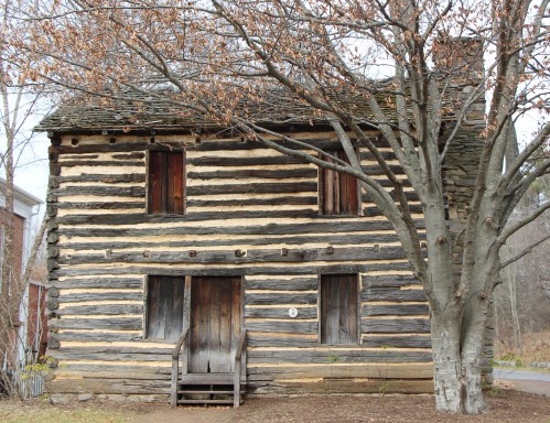 A rustic log cabin with a wooden door, surrounded by a bare tree and a grassy area.