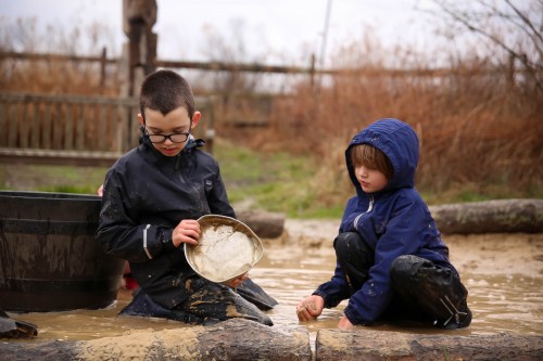 Two children play in a muddy area, one panning for something in a shallow puddle while the other watches intently.