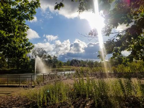 A serene park scene with a fountain, lush greenery, and sunlight filtering through clouds.