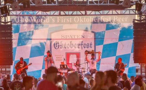 A lively band performs on stage at an Oktoberfest event, with a large banner in the background and an engaged crowd.