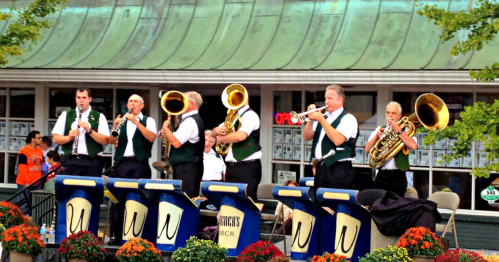 A brass band performs outdoors, featuring musicians with various instruments and colorful flower arrangements in front.