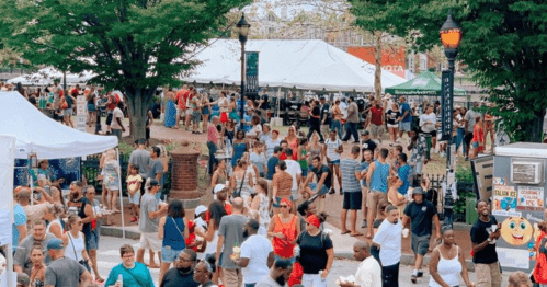 A bustling outdoor festival scene with crowds, tents, and food vendors under trees on a sunny day.