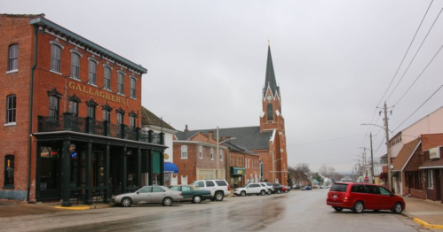 A quaint street scene featuring historic buildings, a church steeple, and parked cars on a cloudy day.