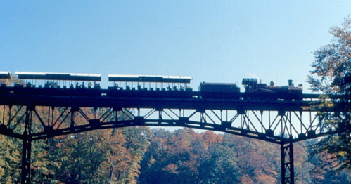 A vintage train crosses a tall bridge, silhouetted against a clear blue sky and colorful autumn trees.