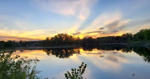 A serene lake at sunset, reflecting colorful clouds and trees along the shore.