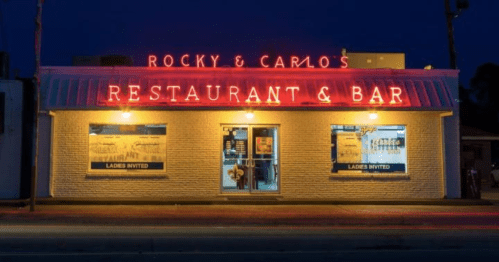Neon-lit exterior of Rocky & Carlo's Restaurant and Bar at night, featuring large windows and inviting signage.