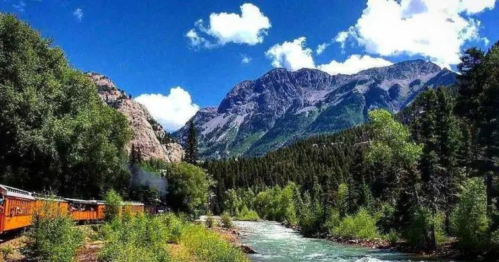 A scenic view of a train along a river, surrounded by lush greenery and majestic mountains under a blue sky.