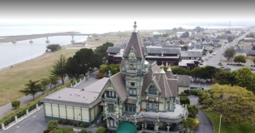 Aerial view of a large, ornate Victorian-style mansion near a waterfront, surrounded by greenery and buildings.