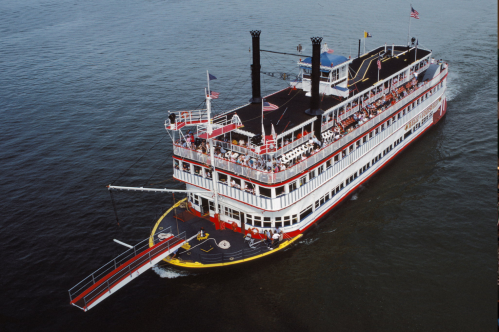 A large riverboat with two smokestacks sails on calm waters, with passengers visible on the deck.