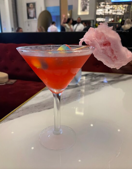 A vibrant cocktail in a martini glass, topped with pink cotton candy, sits on a marble table in a stylish setting.