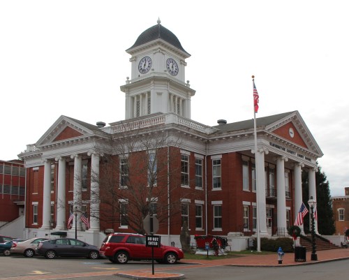 Historic brick courthouse with a clock tower, columns, and American flags, set against a cloudy sky.