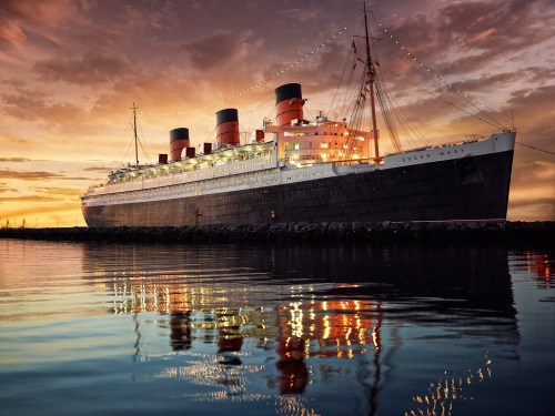 The Queen Mary ship docked at sunset, reflecting in calm waters with colorful lights and a dramatic sky.