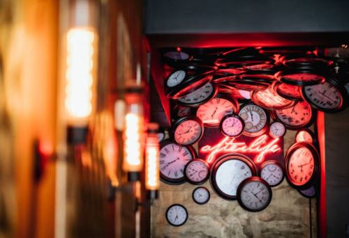 A wall covered in various clocks with a glowing "afterlife" neon sign in red.