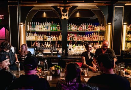 A lively bar scene with patrons at the counter, bartenders mixing drinks, and shelves lined with various bottles.