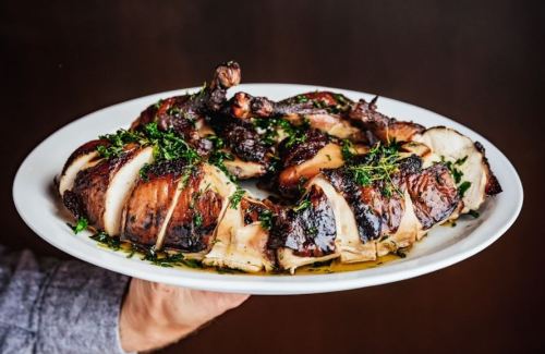 A hand holding a platter of grilled chicken pieces garnished with herbs and drizzled with sauce.