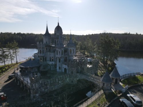Aerial view of a large, ornate castle surrounded by trees and a lake, with a clear blue sky above.