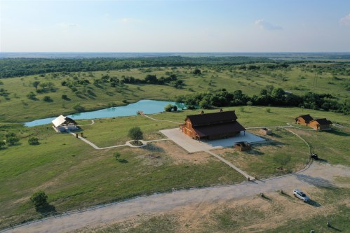 Aerial view of a spacious rural property featuring a large barn, a pond, and green fields under a clear sky.
