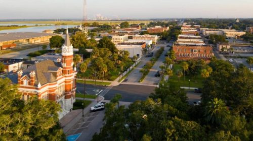 Aerial view of a small town with historic buildings, palm trees, and a clock tower, set against a sunset backdrop.