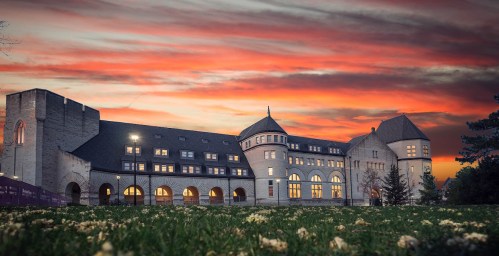 Historic building at sunset, featuring arched windows and a tower, surrounded by green grass and blooming flowers.