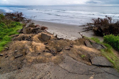 Eroded shoreline with damaged road and debris, overlooking a beach and ocean under a cloudy sky.